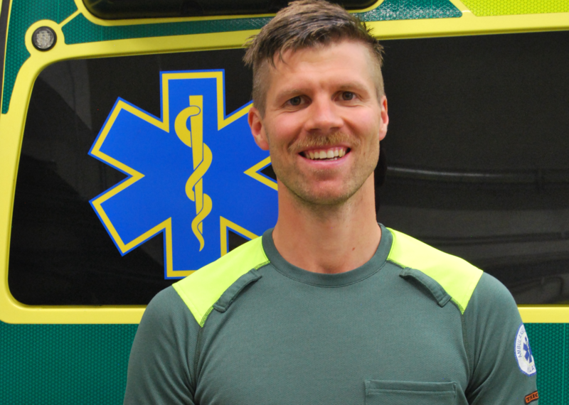 Kristoffer Wibring, forskare och ambulanssjuksköterska som står och ler i ambulansuniform framför en ambulans.
